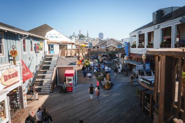 Bustling scene at Pier 39 in San Francisco. Wooden boardwalk lined with shops and eateries. People strolling in the sunny weather, city skyline in the background. clipart