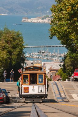 Classic San Francisco cable car with a number 4, ascending a steep hill. View includes marina with yachts, calm bay waters, cityscape, and tourists enjoying a sunny day. clipart