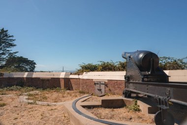 Metal bir taşıma aracındaki eski bir top kumlu zeminde, seyrek bitki örtüsü, tuğla bir duvar ve su manzaralı. Muhtemelen San Francisco 'da tarihi bir yerde..