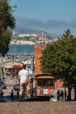 Klasik San Francisco sahne tramvayı dik sokağa tırmanıyor, kondüktör güvertede, Bay View 'a hayran olan adam, arka planda Alcatraz. Körfez kenarında güneşli bir gün..