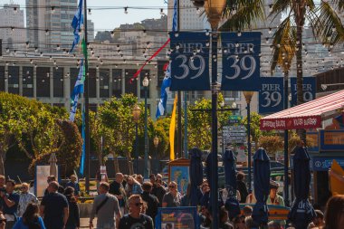 San Francisco 39 numaralı iskelede hareketli bir sahne. Kalabalığın üzerindeki mavi ve altın işaretlere, ziyaretçilerin, palmiye ağaçlarının, şehir mimarisinin, bayrakların, güneşli günün atmosferine odaklan.