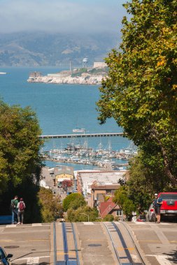 Scenic view of iconic San Francisco hill with cable car tracks, parked cars, green trees. Waterfront with boats near Pier 39. Alcatraz Island in background under hazy sky. clipart