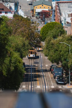 Iconic San Francisco teleferiği park halindeki arabalarla ağaçlık bir caddede ilerliyor. Yükselen manzara tepe araziyi, şehir mimarisini ve mavi gökyüzünü yakalar.