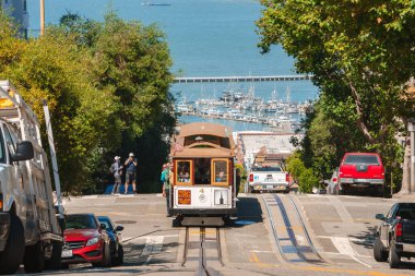Iconic San Francisco scene cable car on steep hill, by bay and marina. Tourists, cars, pedestrians in urban setting with greenery under clear sky. Good for tourism, travel blogs. clipart
