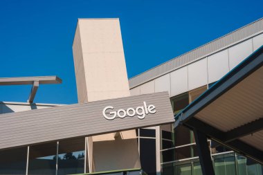 Temiz hatlar, metal ve beton malzemeler içeren cephede Google logosu sergilenen modern bina. Açık mavi gökyüzü çağdaş yüksek teknoloji hissine katkı sağlıyor. Muhtemelen Google ofis kampüsü.