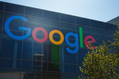 İkonik Google logosuna sahip modern bina açık gökyüzü altında mavi, kırmızı, sarı ve yeşilliklerle övünüyor. Ön planda ağaç var. Cam cephede yansımalar. Google ofisinde yer almaktadır.