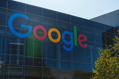 Canlı cam cepheli modern bina renkli Google logosu sergiliyor. Büyük ihtimalle şirket binası. Açık mavi gökyüzü ve ağaç çerçevede. Konum belirtilmedi.