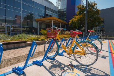 Asfaltlı alanda mavi bisiklet parkında kırmızı sepetli parlak sarı bisiklet. Arka planda modern bina, güneşli hava, yemyeşil.