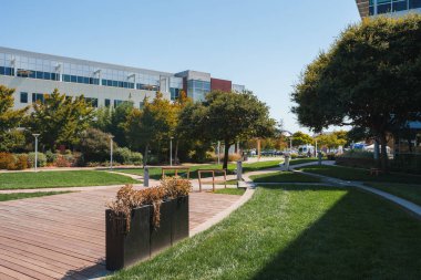 Ahşap kaldırım, metal ekici, çimenlik alan, ağaçlar, lamba direkleri, modern binalar ve kurumsal bir çevreyi işaret eden sokak manzaralı iyi korunan bir park. Huzurlu kentsel ortam.