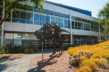 Modern binaların önünde bitkilerle çevrili gerçek boyutta metal T-Rex heykeli. Mavi gökyüzü güneşli bir gün, konum bilgisi yok.
