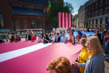 Riga, Letonya 'da coşkulu bir sahne. Büyük bir kalabalık kutlama yapıyor. Odaklanan Letonya bayrağı, arka planda AB bayrakları. Tarihi mimariyle şenlikli bir atmosfer..