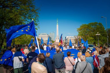 Letonya 'nın Riga kentinde 20. yıldönümünü kutlayan AB ve Letonya bayraklarıyla canlı bir toplantı. İkonik Özgürlük Anıtı arka planda dimdik duruyor. Gururla birleşmiş kalabalık.