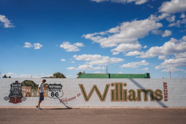Sunny day in Williams, Arizona near Grand Canyon. Person walking by mural celebrating Route 66 connection. Vibrant colors, clear sky, inviting atmosphere. clipart