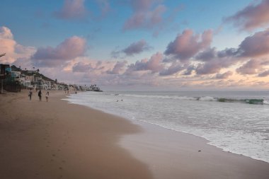 Kaliforniya sahil şeridinde sakin bir sahil manzarası, muhtemelen 17 Mile Drive. Yumuşak ışıklandırma, pastel gökyüzü, nazik dalgalar, su aktivitelerinden hoşlanan insanlar, resimli kıyı manzarası.