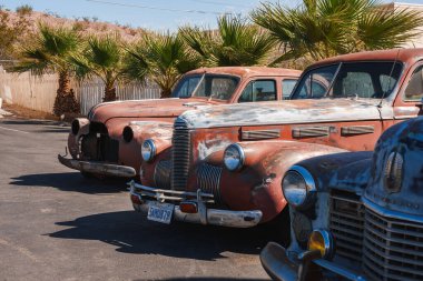 Vintage cars parked in a row, showcasing a rustic appearance reminiscent of mid 20th century American design. Located in Barstow, along Route 66, under a clear blue sky. clipart