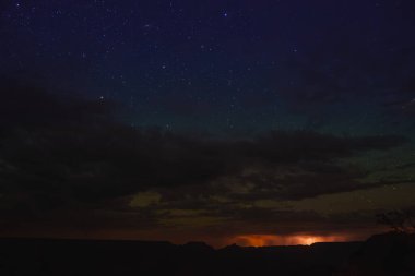 Yıldızlı dramatik gece gökyüzü, siluetli ufuk, uzak kentsel alanı ya da olayı işaret eden ılık parıltı. Bulutlar derinlik ekler. Uzak konum, serbest ışık kirliliği.