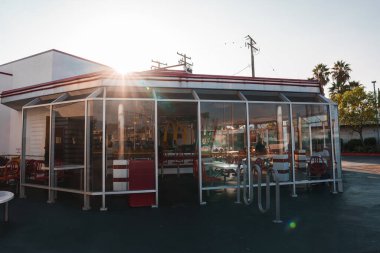 Classic American diner in Los Angeles with mid 20th century style, white exterior, red trim, traditional booth seating, and outdoor area with red and white chairs under a canopy. clipart