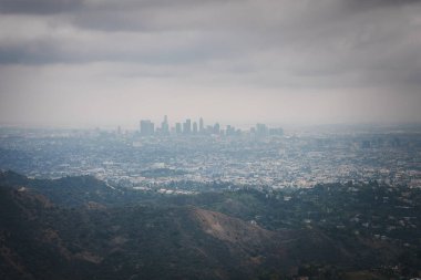 Bulutlu gökyüzü altındaki geniş şehir manzarası, Los Angeles şehir merkezi ufuk çizgisi. Yuvarlanan tepeler ön planda, muhtemelen Hollywood Tepeleri, yumuşak ışıklandırmalı yoğun binalar. Yüksek görüş açısı yakalama.