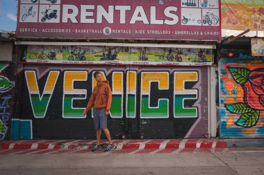 Venedik Plajı, Los Angeles 'ta renkli bir mağazanın önünde grafiti tarzı sanat eserleriyle yürüyen bir adam. Dükkanda kiralar, hizmetler ve aksesuarlar sunuluyor. Sahil kenarında rahat bir hava..