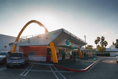 McDonalds restaurant in Los Angeles features iconic golden arches, mid century modern style, red and yellow color scheme, operational with cars in the parking lot, under a warm sunset sky. clipart