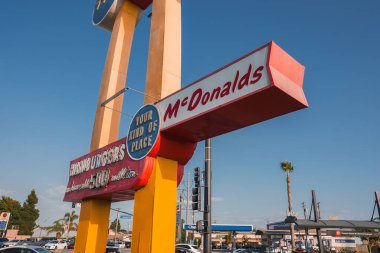 Iconic McDonalds canlı bir şehir ortamında açık mavi gökyüzüne karşı imza atıyor, muhtemelen Los Angeles 'ta. Klasik altın kemer logosu ve kırmızı arkaplan ile klasik görünüm.