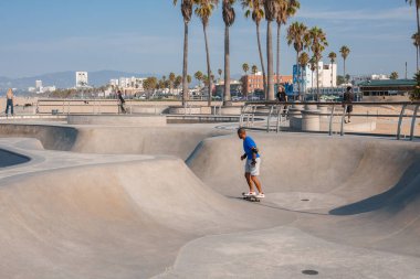 Venice Beach, Los Angeles 'ta güneşli bir gün. Hareket eden patenciler, palmiye ağaçları, açık mavi gökyüzü, sakin bir atmosfer. Kaykay yetenekleri için ideal..
