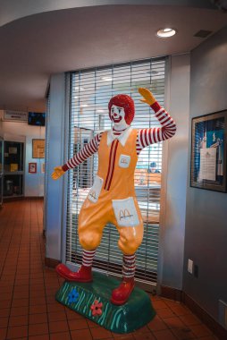 Life size Ronald McDonald statue in iconic attire, waving and welcoming. Indoors with blinds, likely in Los Angeles. Background hints at restaurant setting. clipart