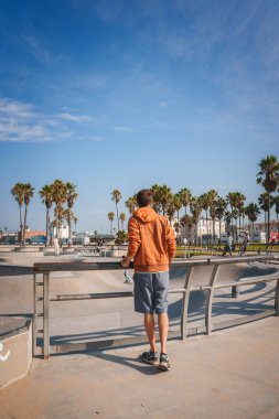 Paten parkının kenarında, turuncu kapüşonlu, gri şortlu, Venice Beach, LA 'de spor ayakkabısı olan manzaralı bir adam. Güneşli bir gün, palmiye ağaçları, arka planda patenciler.