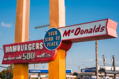 Büyük altın kemerler, kırmızı HAMBURGERS ve 500 milyondan fazla satılan metin içeren klasik McDonalds tabelaları. Kentsel ortamda palmiye ağacı ve mavi gökyüzü olan Americana atmosferi, muhtemelen Los Angeles, California.