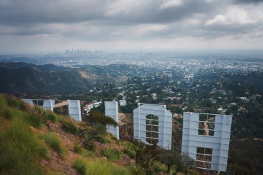 Iconic Hollywood Sign view from the hills of Los Angeles with sprawling cityscape in the distance. Overcast sky, rugged terrain, and downtown skyline visible. clipart