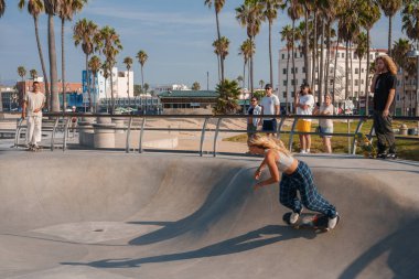 Venice Beach, Los Angeles 'ta canlı kaykay sahnesi. Beyaz atletli ve mavi ekose pantolonlu bir kadın beton bir kasenin içinde, palmiye ağaçları ve izleyicilerle çevrili. Güneşli bir gün..