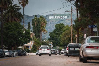 Los Angeles sokak manzaralı ve arka planda Hollywood tabelası var. Park edilmiş arabalar, yeşil ağaçlar, bulutlu gökyüzü, palmiye ağaçları, yayalar yok. Hollywood dönüm noktası.