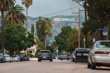 Park halindeki arabalarla, palmiyelerle ve tepelerle dolu bir caddeye bakan ikonik Hollywood tabelası. Bulutlu gökyüzü havayı artırır. Zemin kat perspektifi. Muhtemelen Los Angeles 'ta..