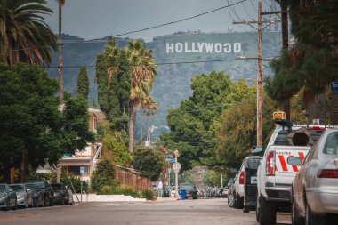 Arka planda ikonik Hollywood tabelası, park halindeki arabaların, palmiye ağaçlarının ve yayaların olduğu Los Angeles sokak sahnesi, rahat Güney Kaliforniya atmosferini yakalıyor..