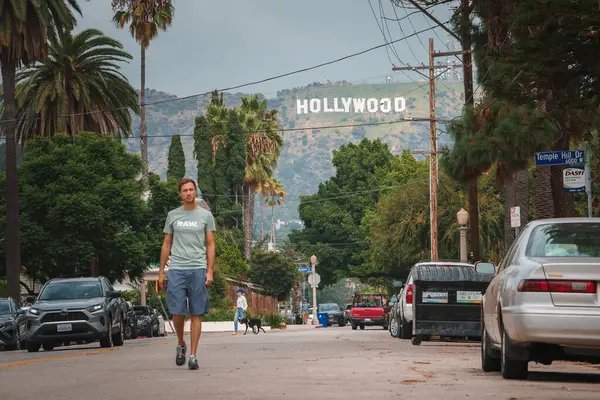 Los Angeles caddesinde park etmiş arabalarla, palmiye ağaçlarıyla dolaşan bir adam. Hollywood tabelası görünür, bulutlu gökyüzü. Günlük giysiler, rahat yürüyüş. Hollywood mahallesi heyecanı.