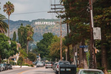 Los Angeles tepelerinde Hollywood tabelası olan manzaralı sokak manzarası. Park edilmiş arabalar, konut hissi, palmiye ağaçları, bulutlu gökyüzü. Beachwood Dr. Kavşak.