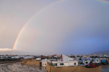 Sprawling desert settlement with wet ground and rainbow sky. Mix of tents, RVs, and structures. Transient city amidst barren landscape. Mountain range in distance. clipart