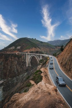 Scenic coastal road winds along rugged cliffside in coastal California, USA. Concrete bridge spans deep canyon with vehicles for scale, under clear sky. clipart