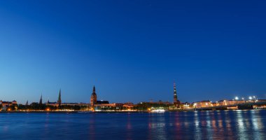 Riga, Letonya 'da sakin bir akşam Riga Katedrali ve St. Peters Kilisesi' nin aydınlatılmış ufuk çizgisini gösteriyor. Sıcak ışıklar Daugava Nehri sularından yansıyor.