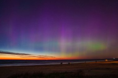 Letonya 'daki Jurmala plajı üzerinde canlı mor, yeşil ve mavi tonlarıyla büyüleyici bir aurora borealis görüntüsü. Sakin deniz renkleri yansıtır, sahneyi güçlendirir..