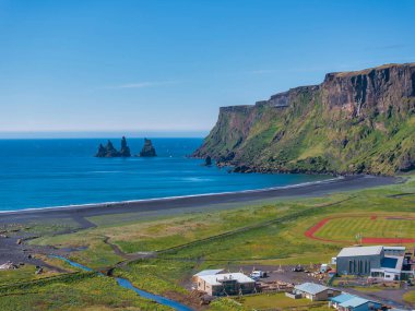 Üç deniz yığını, yeşil çimenler ve ön planda akan küçük bir nehrin yer aldığı İzlanda 'daki siyah kum plajının çarpıcı hava manzarası..