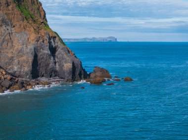 Yeşil bitki örtüsü ve kahverengi kayayla kaplı kayalık bir uçurumun havadan görünüşü, açık mavi okyanusa küçük kayalar ve adalar ile dalgalanıyor..