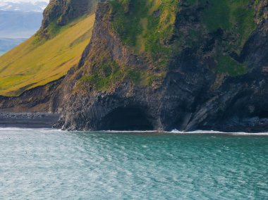 İzlanda 'nın siyah kumsal manzarasında bazalt sütunlardan yapılmış kayalık bir yüzün içinde büyük bir mağara var. Derin mavi su ve açık mavi gökyüzü sahneyi tamamlıyor..