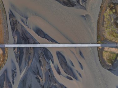 İzlanda 'da açık kahverengi bir nehir boyunca uzanan basit bir köprünün kuş bakışı görüntüsü. Çevresindeki manzara, doğal güzelliği vurgulayan kahverengi ve yeşil renklerin bir karışımını içerir..