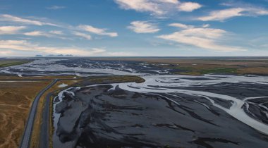 İzlanda 'da siyah kumun içinden geçen geniş ve sığ bir nehrin çarpıcı hava manzarası. Sol tarafta mavi gökyüzü ve beyaz bulutlarla dolu bir yol var..