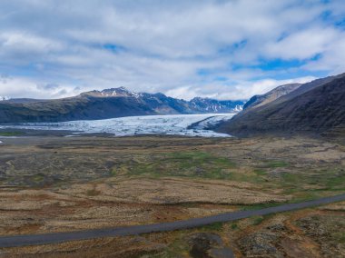 İzlanda 'da engebeli dağlarla çevrili bir buzulun çarpıcı hava manzarası. Ön plandaki bir yol nefes kesici manzaraya ölçek ve perspektif ekler..
