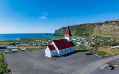 Yeşil tepeler ve dağlarla çevrili küçük bir İzlanda kasabasındaki tepenin üzerinde kırmızı çatılı beyaz bir kilisenin çarpıcı hava manzarası okyanusu ve siyah bir kumsal manzarası..