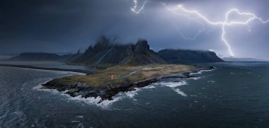 A powerful lightning strike illuminates the rocky cliffs and churning ocean of Icelands coastline, showcasing the raw beauty of Vestrahorn mountaine on Stokksnes cape clipart