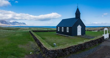 Çimenli bir tarlada beyaz bukleli, dağlarla çevrili ve İzlanda 'nın arka planında bir su kütlesi olan siyah bir kilisenin hava görüntüsü..