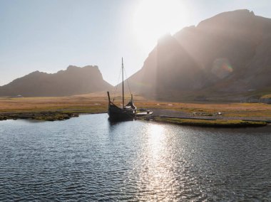 A traditional Viking longboat sits in calm waters under a bright sun, with rugged, snow-capped mountains in the background. Evocative scene of a bygone era.